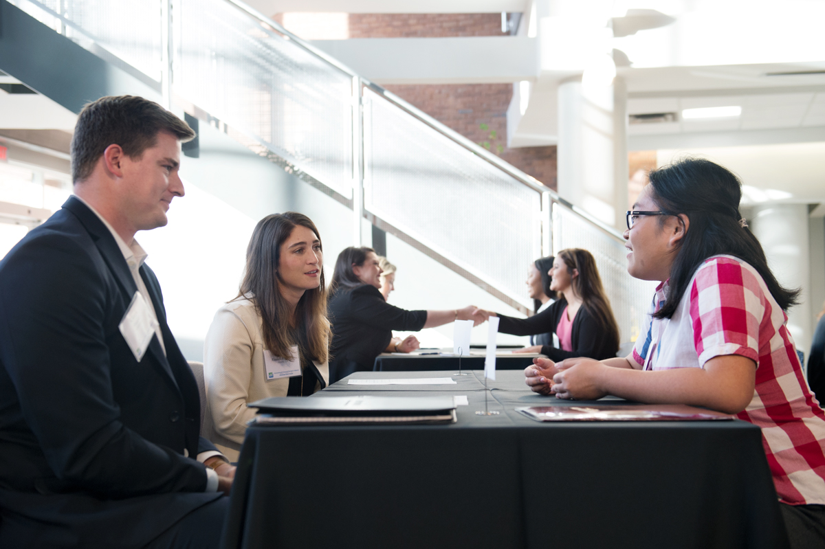 Accounting student talks with two accounting firm reps at a table in the McCool atrium while others shake hands in background.