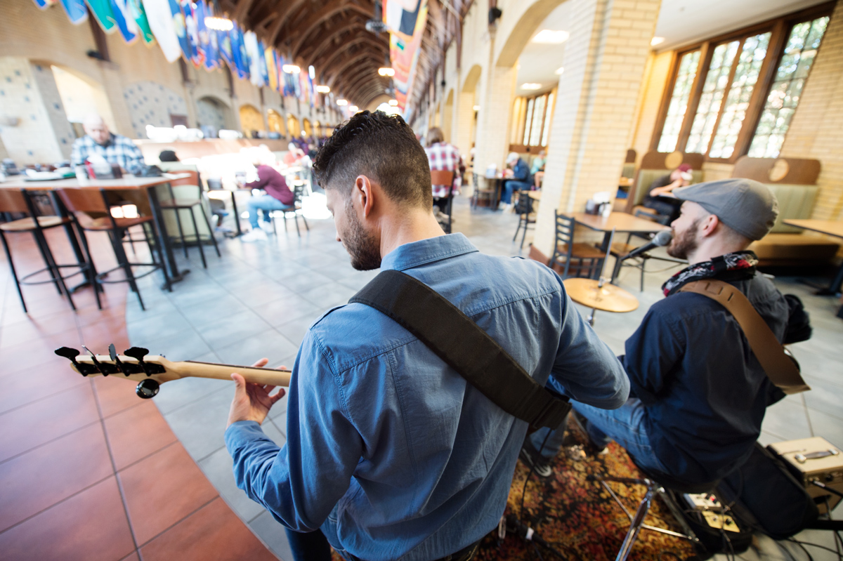 A musical duo plays to lunchtime diners at Perry Cafeteria, with their backs to the camera and Perry&amp;#039;s flags in the background.