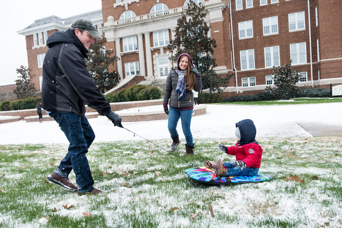 Parents pull their toddler on a boogie board sled in the snowy grass in front of Swalm.