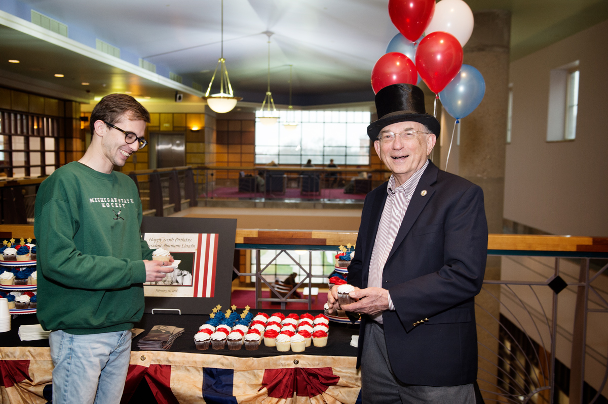 In front of birthday cupcake display in the library, John F. Marszalek wears a Lincoln style tophat with Library student worker.