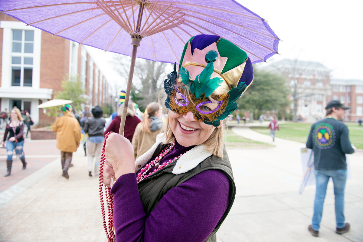 Mardi Gras staff parade participant smiles beneath her umbrella with colorful mask beads and hat.