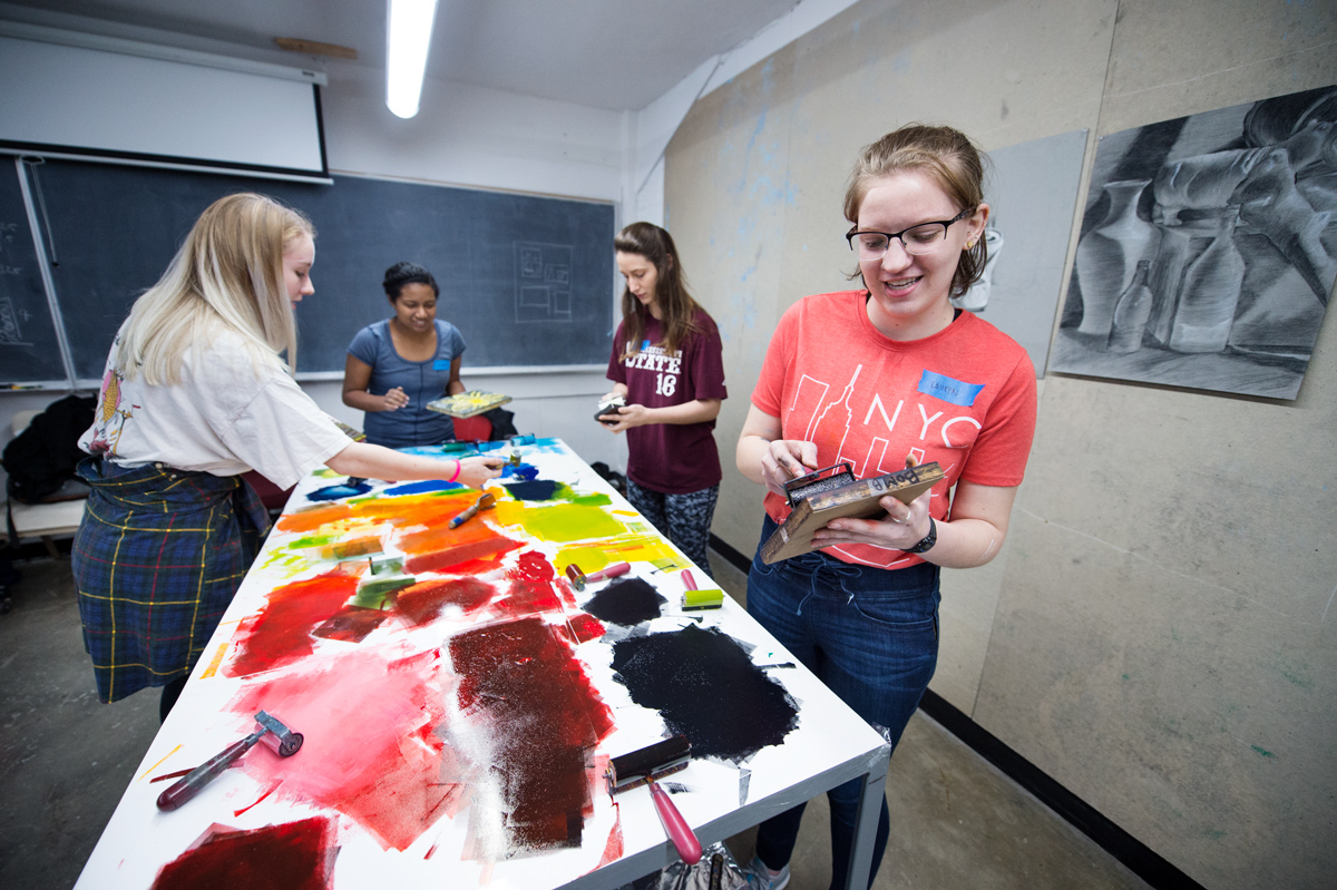 Four art students work around a table covered with colorful patches of ink, using brayers to ready printing blocks. 