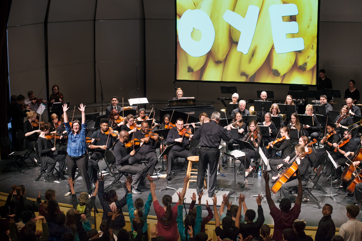 A wide shot of the Starkville-MSU Symphony Orchestra with children in the audience participating with their arms outstretched.