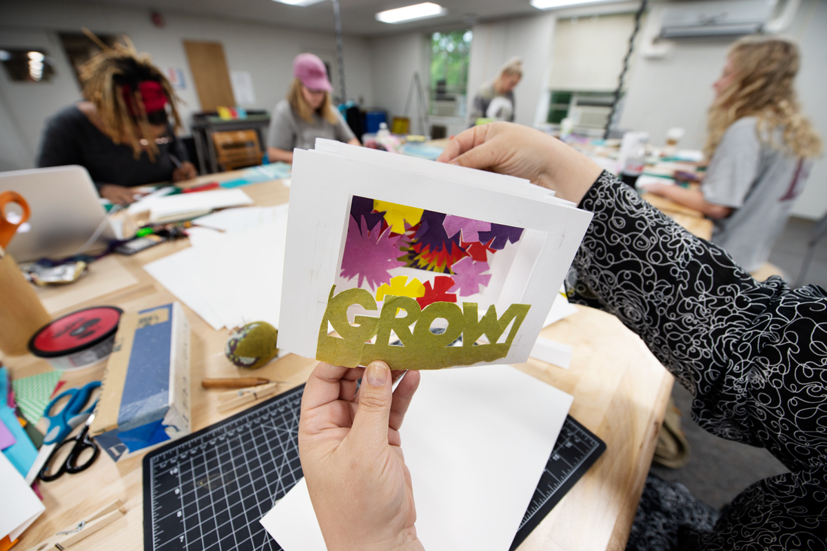 While students work around a table in the background, Professor Powney&amp;#039;s hands hold a tunnel book sample decorated with flowers 