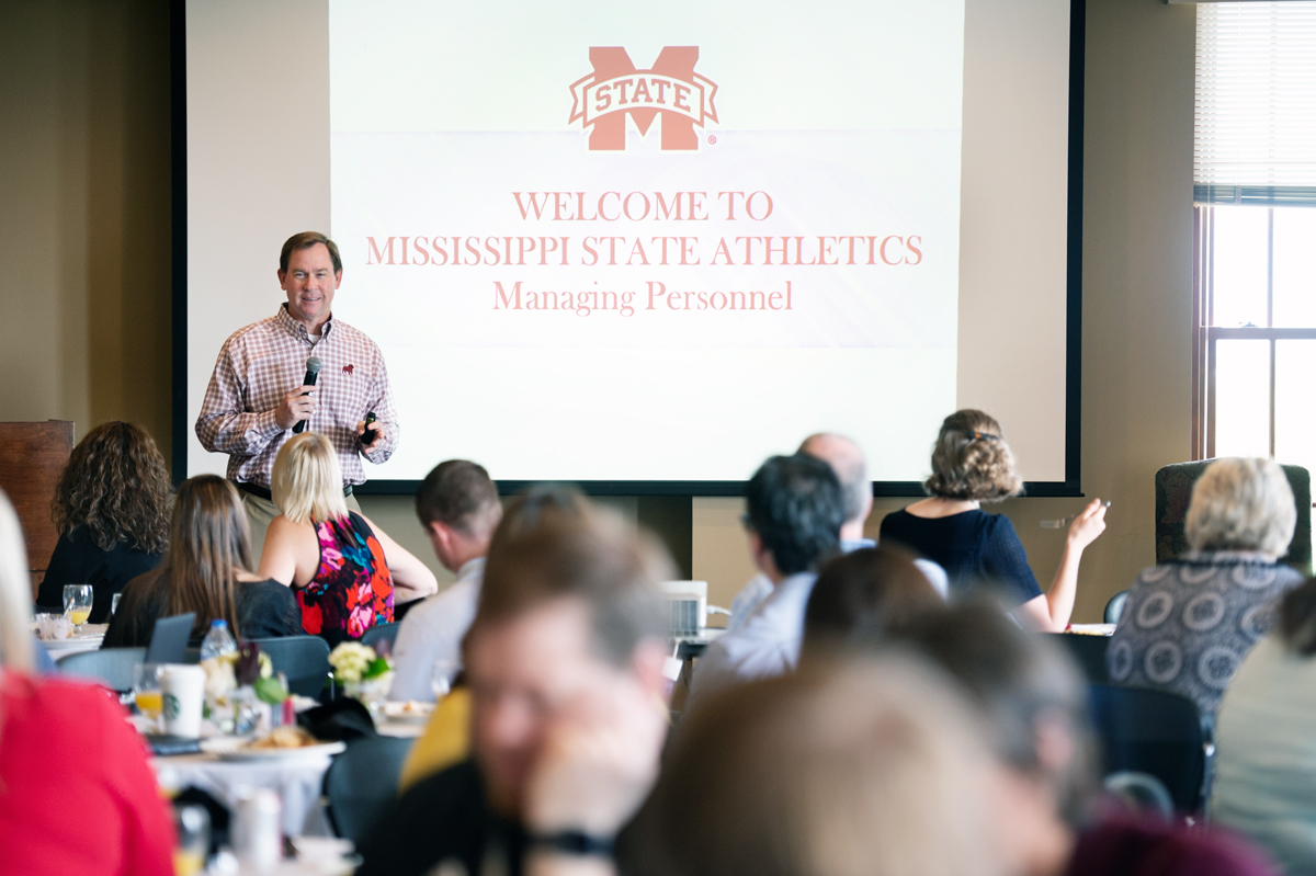 Over a foreground of 2018 SECCA Attendees eating breakfast, MSU Athletic Director John Cohen speaks about managing personel.