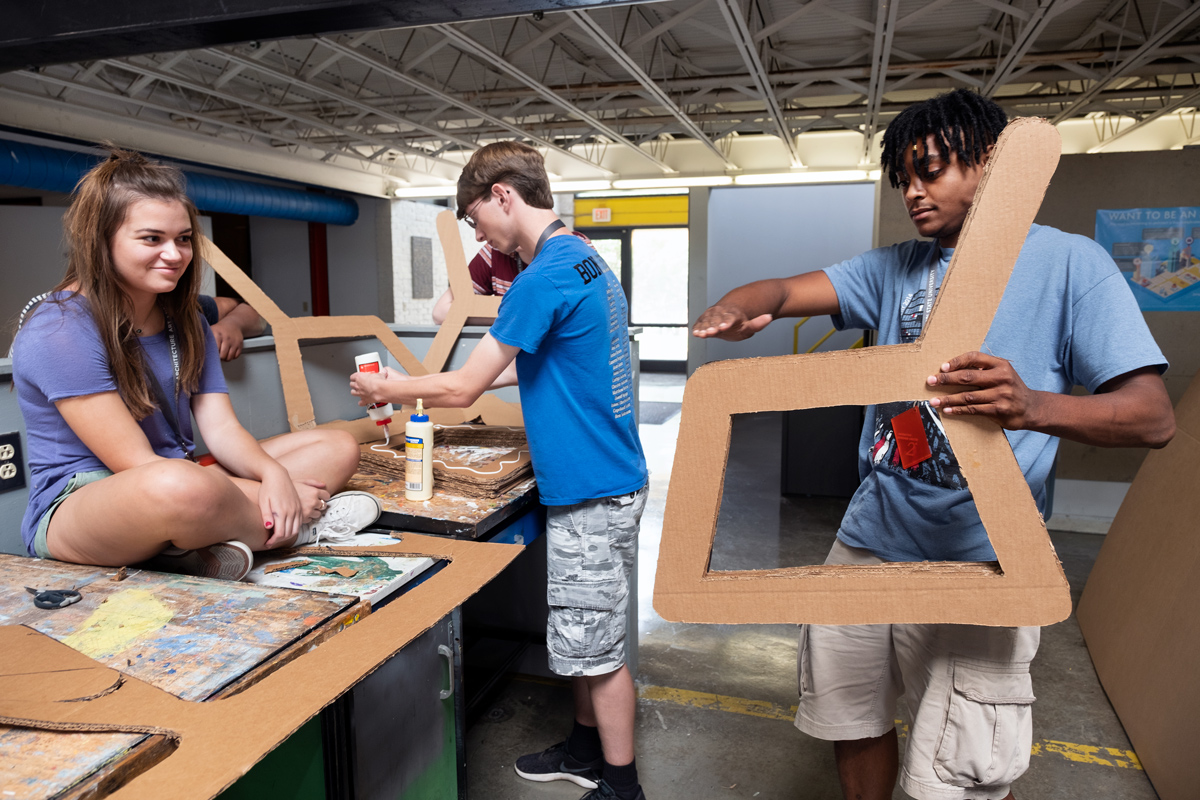 With high school Design Discovery participants looking on, architecture student Jordan Smith holds their cardboard chair project