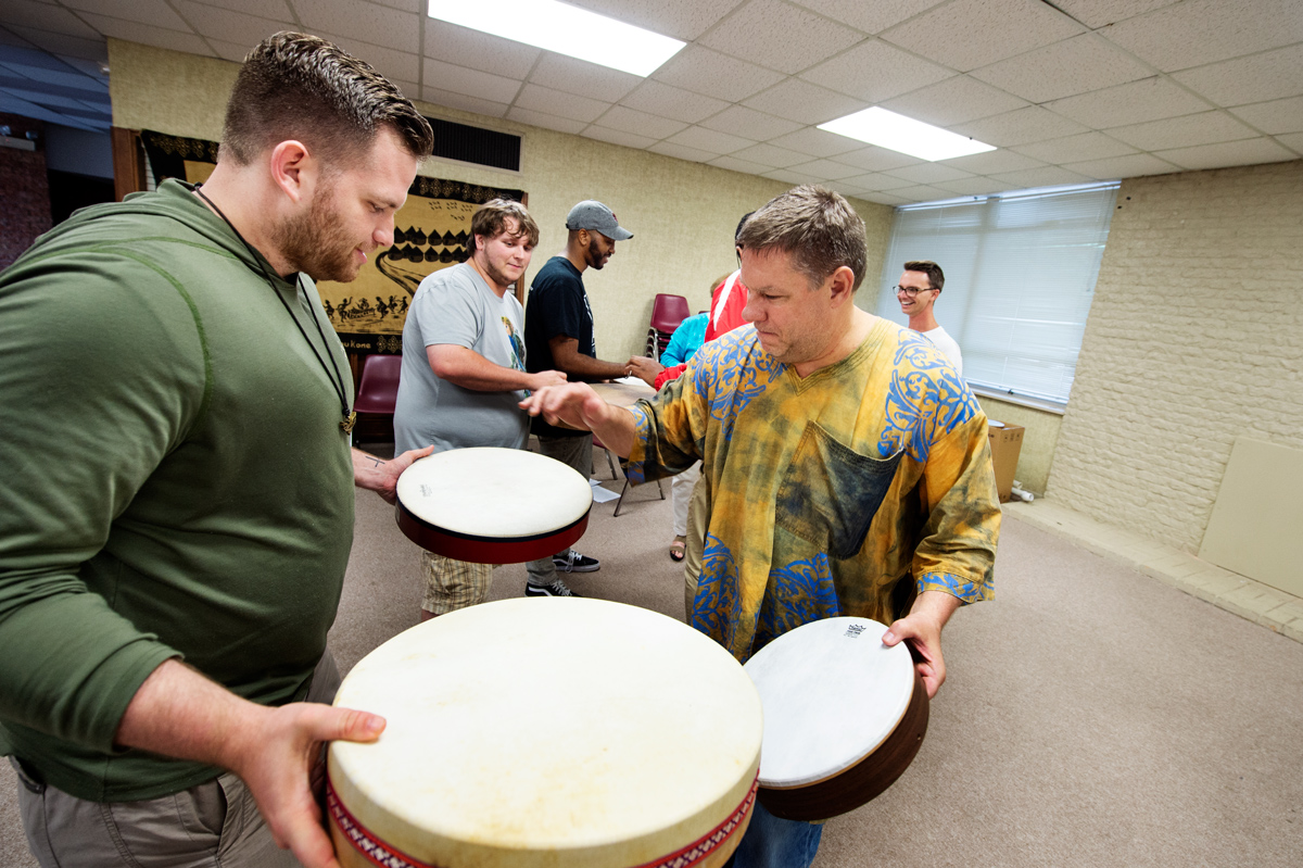 Music Professor Robert Damm drums with a student on hand-held frame drums while other students do the same in the background. 