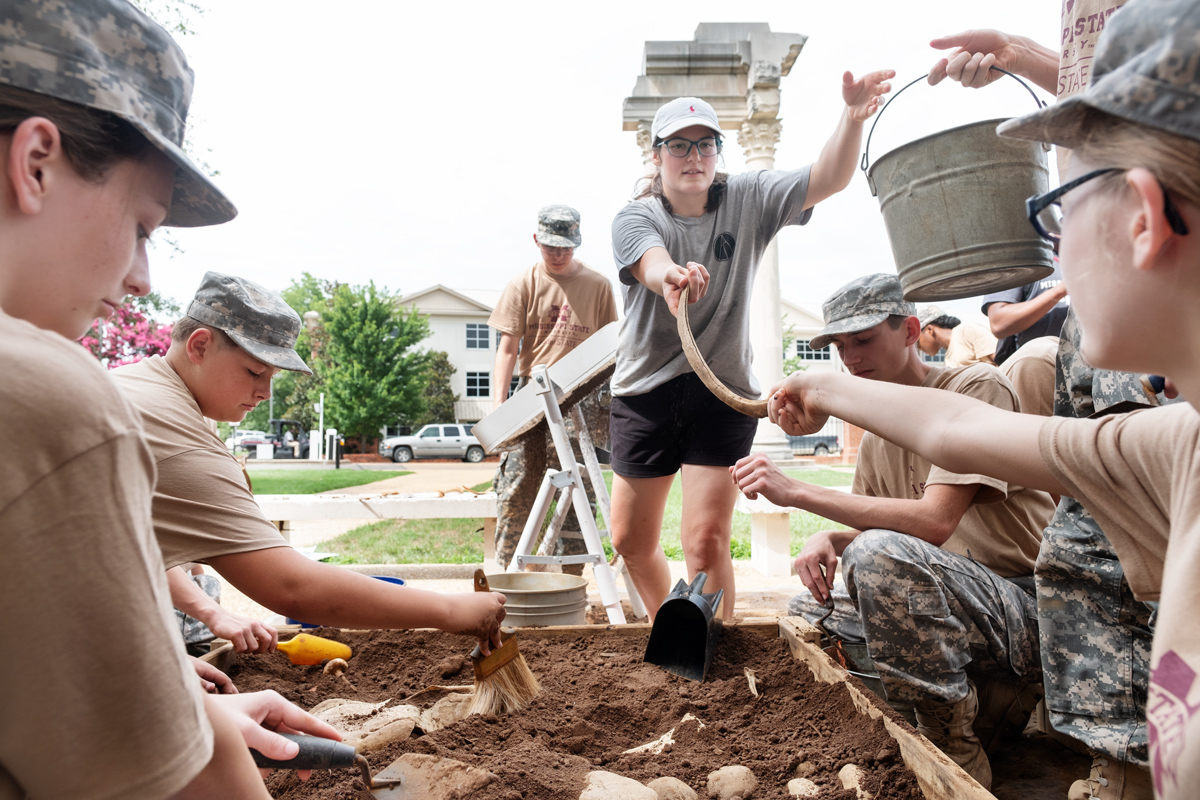 With JROTC Cadets digging archaeological artifacts in the foreground, student Karen Hall hands the team a new bucket.