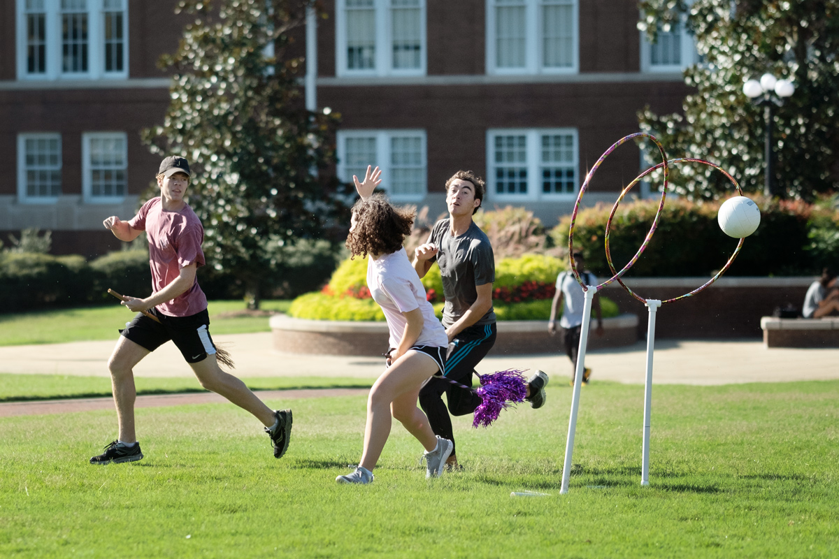 Three students &amp;quot;fly&amp;quot; with brooms around the Drill Field, trying to get the quaffle ball through the goal hoops.
