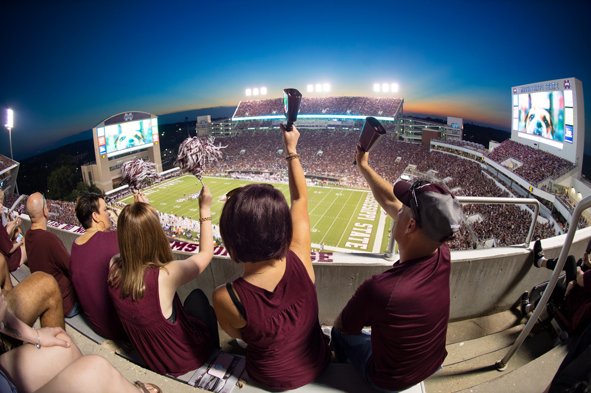 Fans on the top of Davis Wade Stadium ring cowbells and shake pom poms with the bowl of the stadium and sunset beyond them.