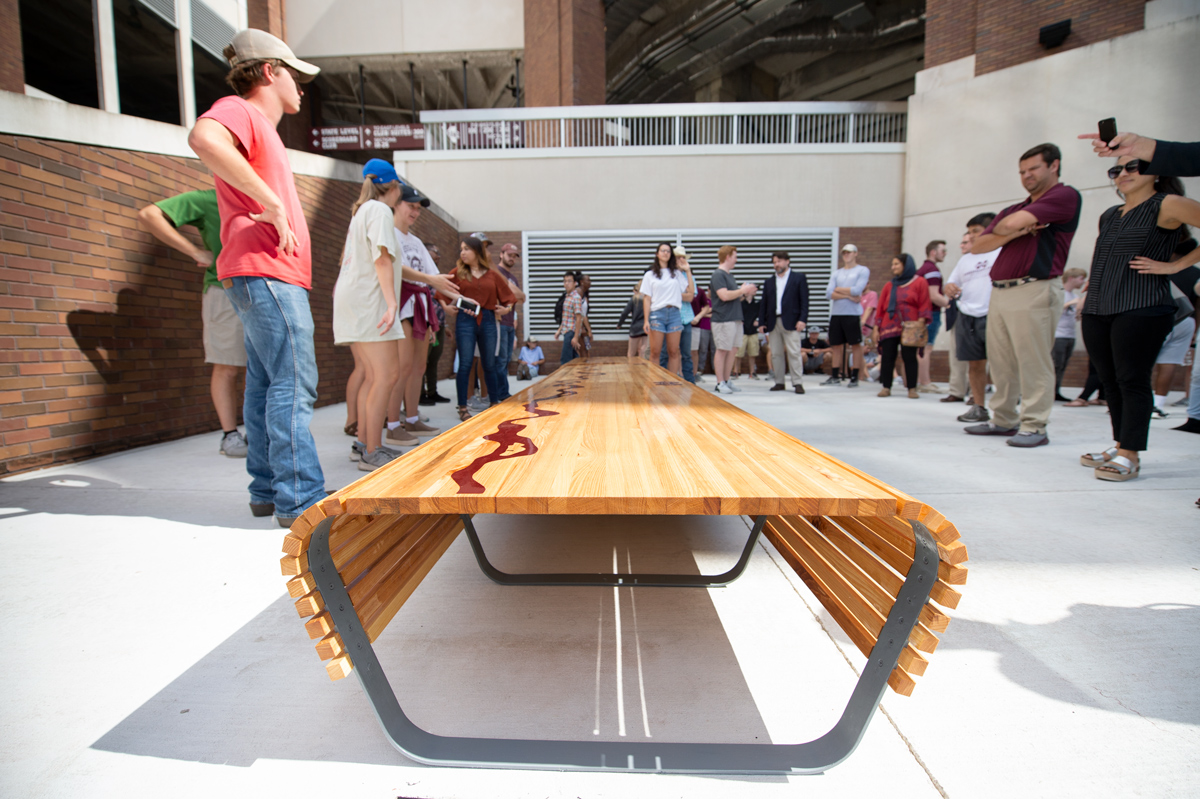 With a long wooden bench in the central foreground and Davis Wade Stadium above, students and professors circle behind. 