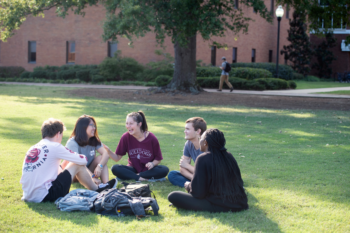 A circle of five undergraduate students share a laugh while sitting on the grass of the Drill Field in between classes.