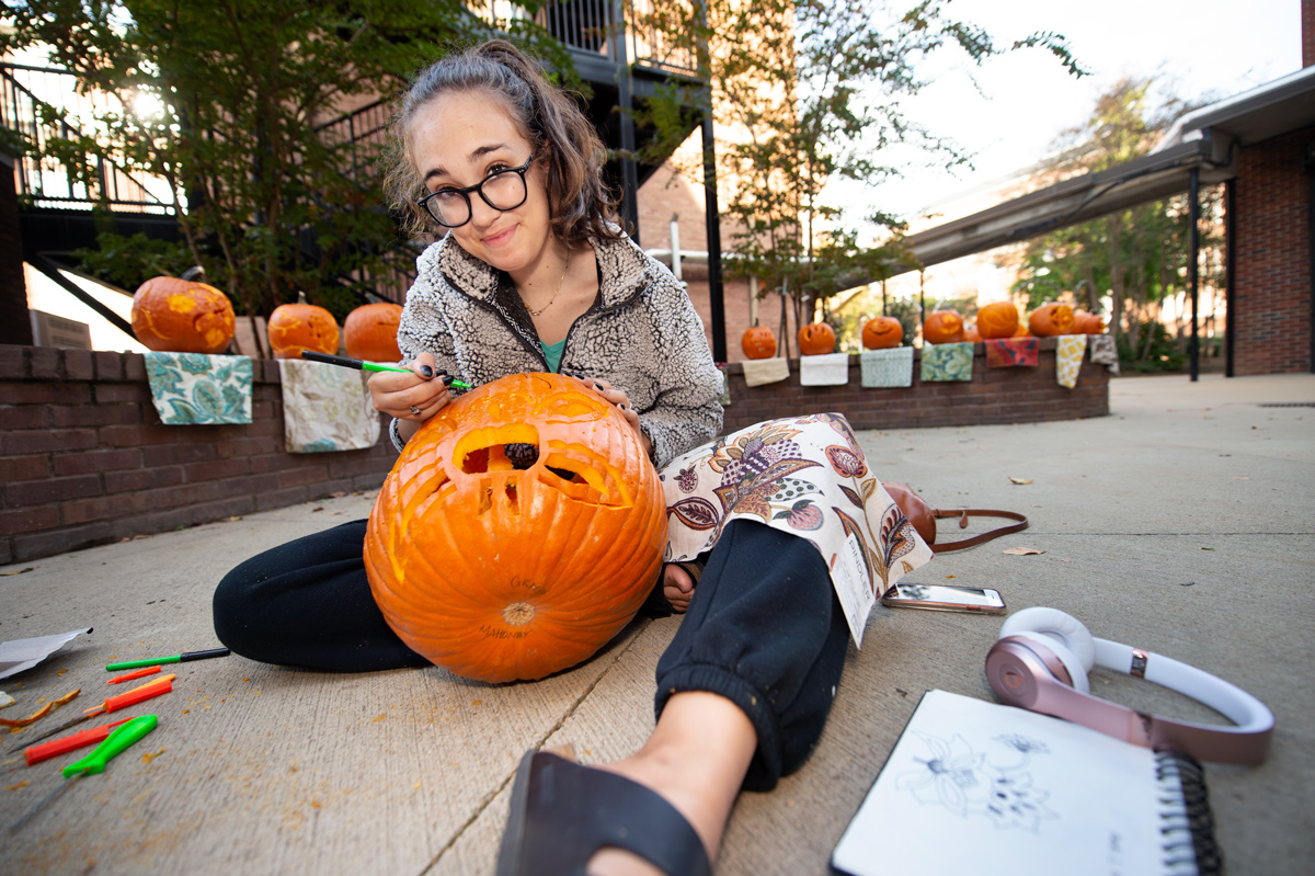 Grace Mahoney carves a pumpkin on her lap with reference textile designs around her and her classmates&amp;#039; pumpkins behind.