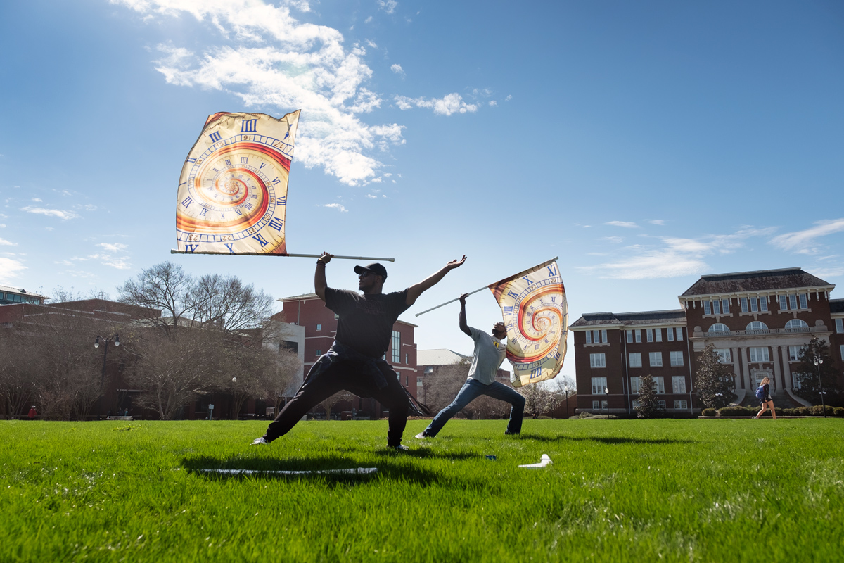 On a sunny on the lush green Drill Field grass, two students twirl clock-themed flags during their color guard practice.