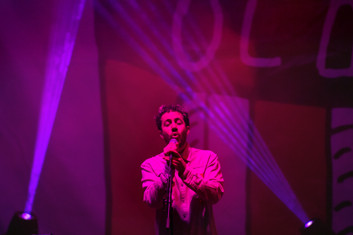 Lewis Del Mar sings into the microphone, lit by fuschia lights with beams of purple lights behind.