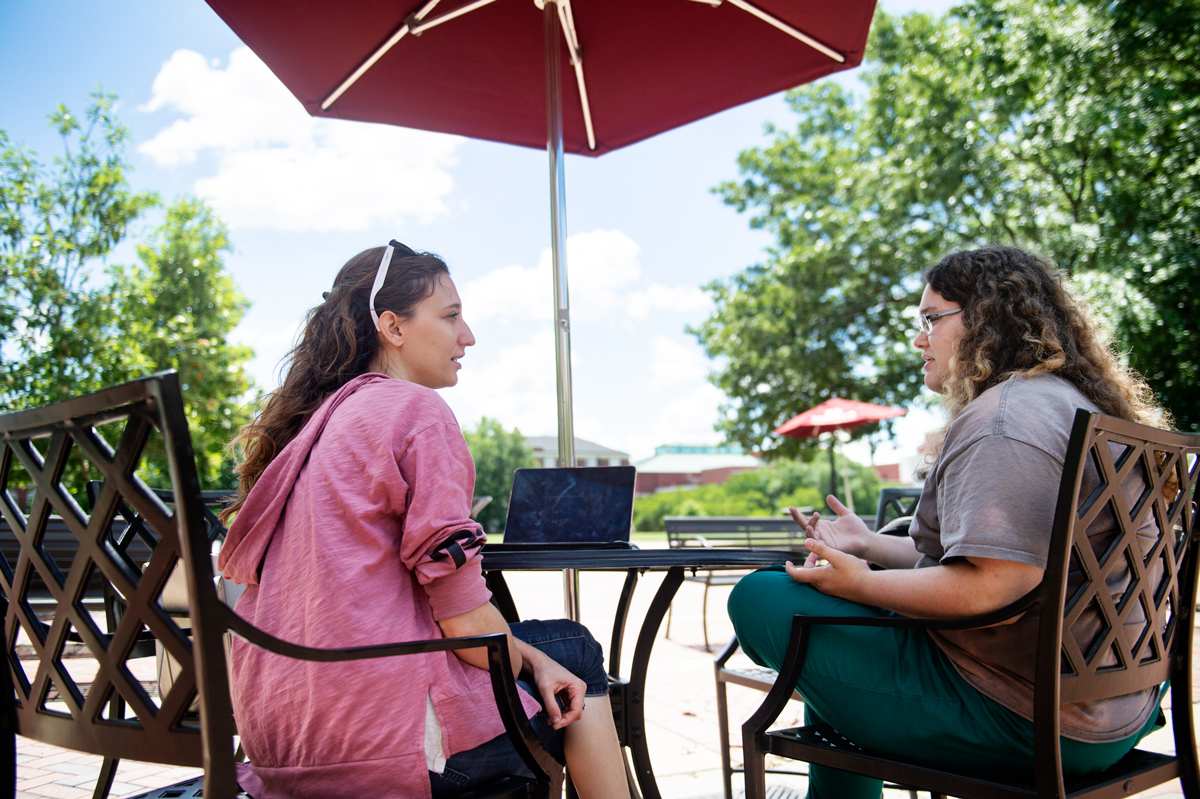 Graduate students Gwyneth Jones and Carolina Baruzzi talk in the shade of an outdoor umbrella with the sky and trees behind. 