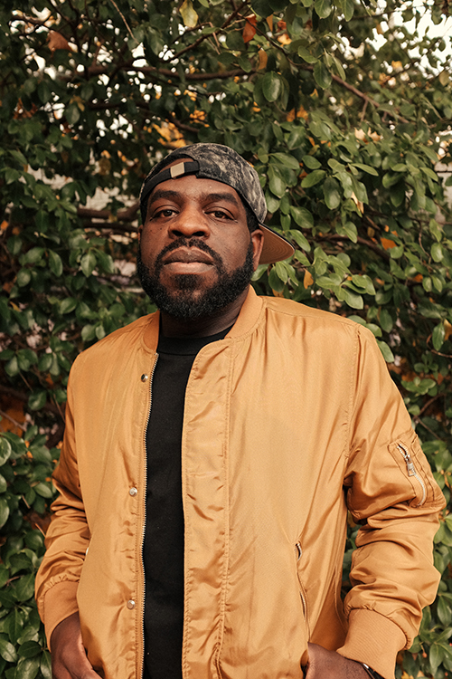 Award-winning author Hanif Abdurraqib stares at the camera while standing in front of trees.