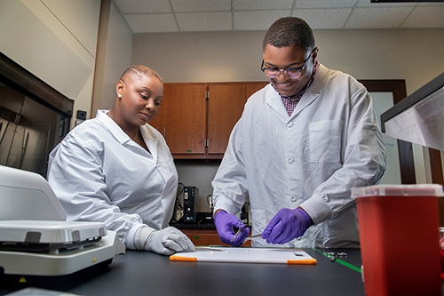 MSU Associate Professor LaShan Simpson looks on as MSU biological engineering student Christopher Robinson uses scissors and tweezers in a lab setting.