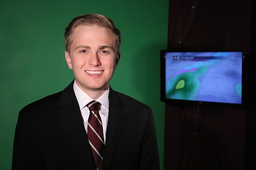 Alex Forbes wears a suit and tie pictured in front of a broadcast green screen and television monitor