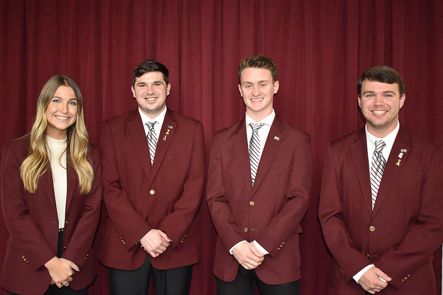 One female student and three male students are smiling and wearing maroon coats in front of a maroon background