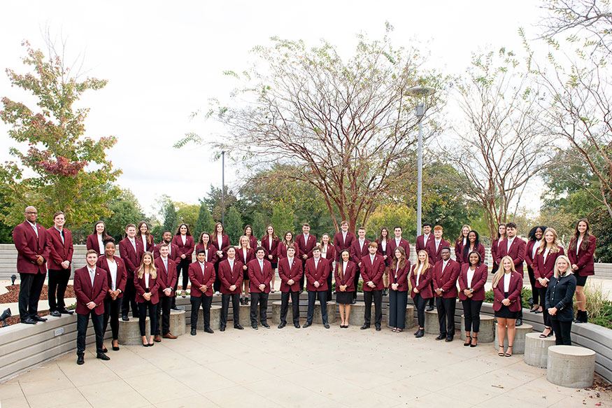 A group of Alumni Delegates wearing maroon coats pose for a photo outdoors. 
