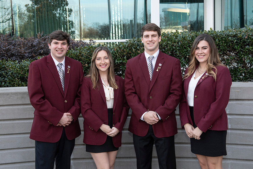 Four officers of the student organization Alumni Delegates stand together wearing maroon blazers