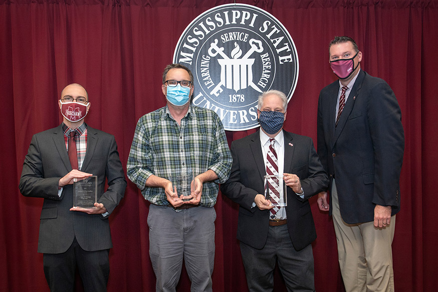 Four men stand (three holding awards) in front of the MSU seal.