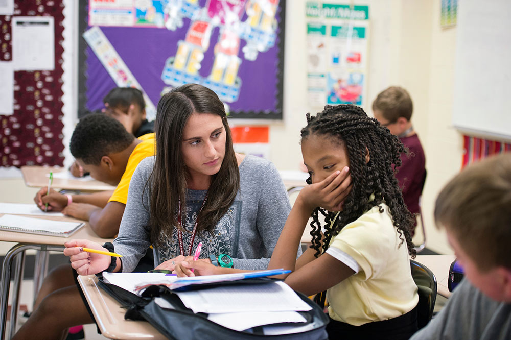 A female college student works with a young girl in a classroom setting.