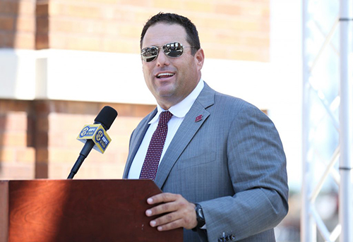MSU Head Baseball Coach Chris Lemonis wears shiny sunglasses and speaks at a podium during a campus press conference.