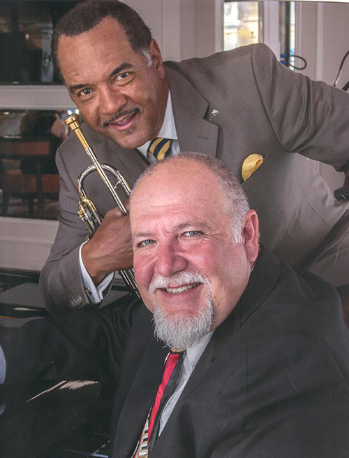 Wendell Brunious holds a trumpet while standing next to Tom Hook, pictured seated at a piano