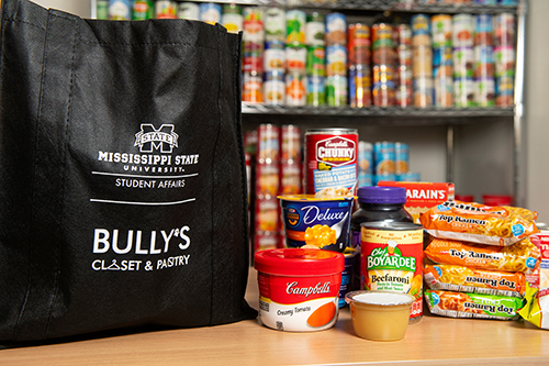 A black tote bag with MSU Student Affairs and Bully’s Closet and Pantry logos is pictured next to soup cans, applesauce, macaroni and cheese, jelly, rice and Ramen noodles