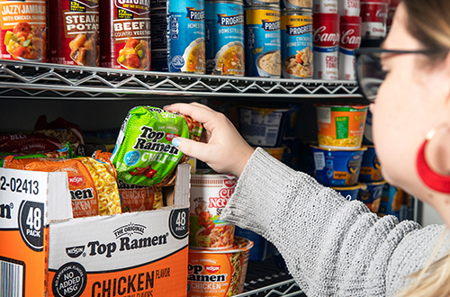 A woman adds a pack of chili-flavored Ramen noodles to a shelf of canned food.