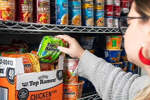 A woman adds a pack of chili-flavored Ramen noodles to a shelf of canned food.