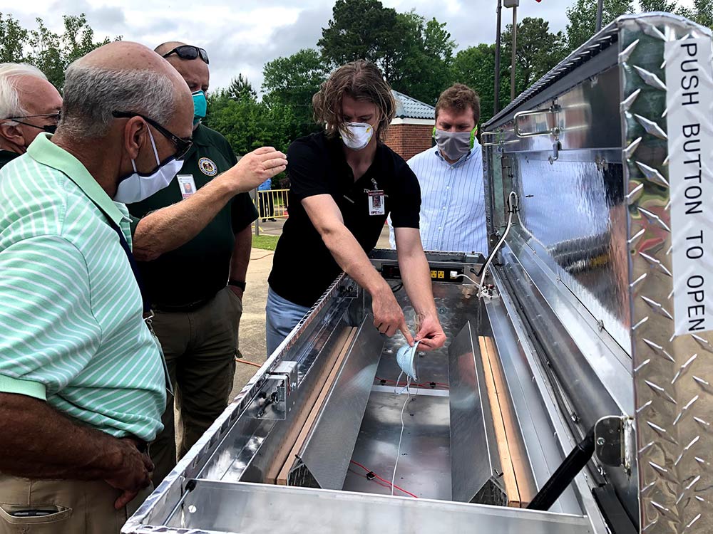 A young man demonstrates how to operate an ultraviolet sterilization device made from a truck toolbox as other men watch.