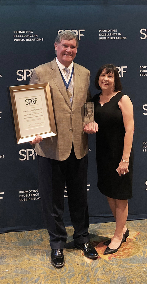 Checky Herrington, joined by his wife Chellie, holds the 2019 Professional Achievement Award certificate in front of a SPRF logo backdrop.
