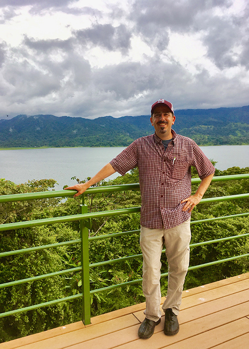 David Hoffman smiles for the camera while standing near a rail with trees, mountains and a body of water in the distance.