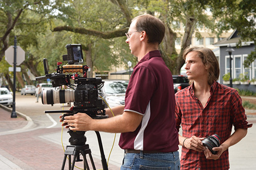 MSU University Television Center Director David Garraway operates a video camera as UTC Senior Producer James Parker looks on while holding a large black camera lens.
