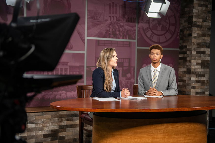 Two students, a young woman and man, are on the set of the “Take 30 News” program with a television camera in the foreground and a stage light overhead and MSU themed set background.