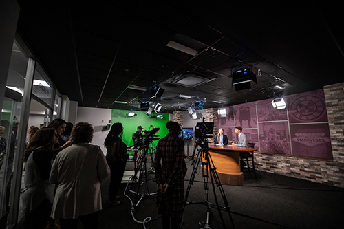 Two student news anchors on set and others working and looking on at the fully operational television studio in Mitchell Memorial Library’s MaxxSouth Digital Media Center.