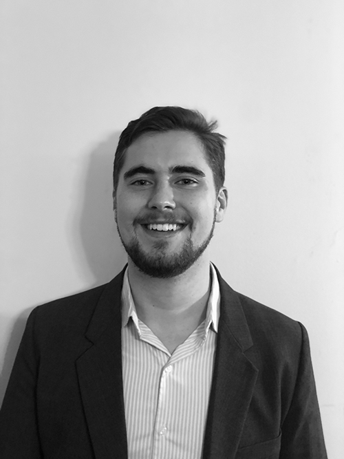 Black-and-white photo of Duncan Thomas smiling while standing in front of a wall.