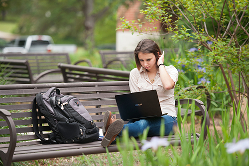 A college-age woman sits on a bench outside looking at her laptop with trees and flowers in the background