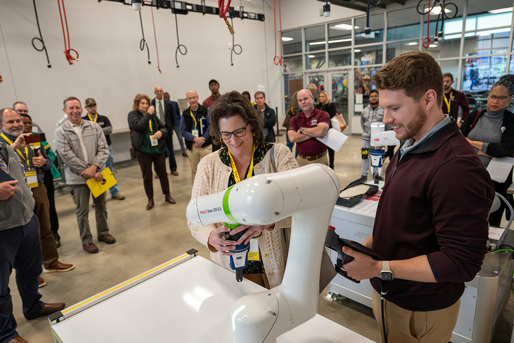 A man demonstrates how to use robotic manufacturing equipment in a lab full of people.