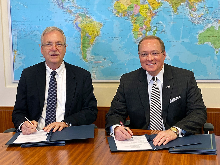 Two men seated at a table holding pens to sign documents. A world map is on the background wall.