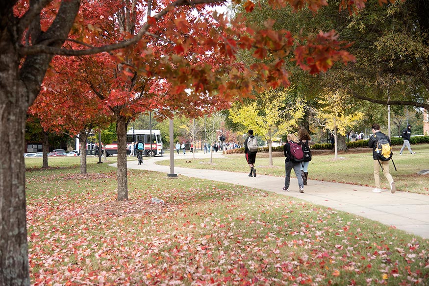 Students walk on a sidewalk as trees are seen throughout the campus landscape
