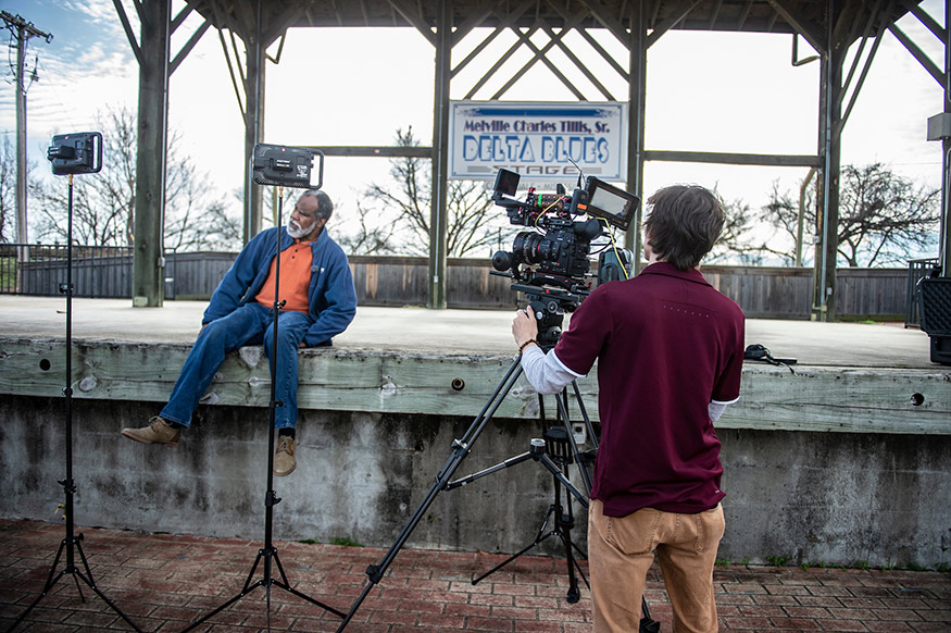 A man points a television camera toward a man sitting in front of a sign that says Delta Blues