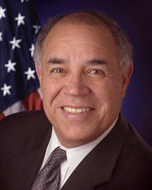 Fred Gregory smiles for the camera while seated in front of the American flag.