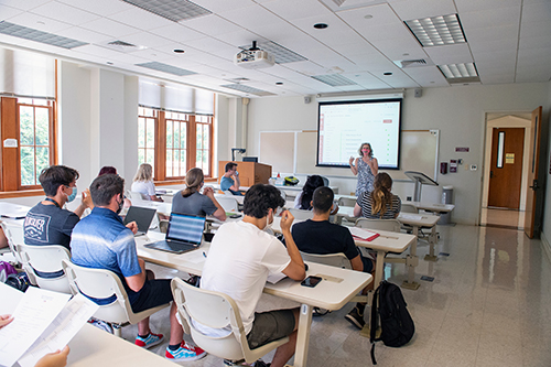A faculty member speaks to college students from the front of a classroom.
