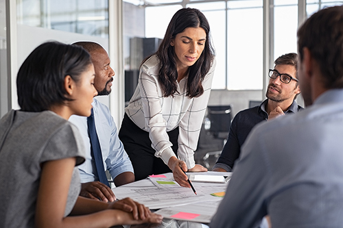 A professional woman points to papers on a conference room table and talks with colleagues 