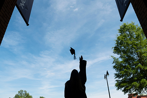 A graduate, silhouetted against a blue sky, tosses a graduation cap in the air