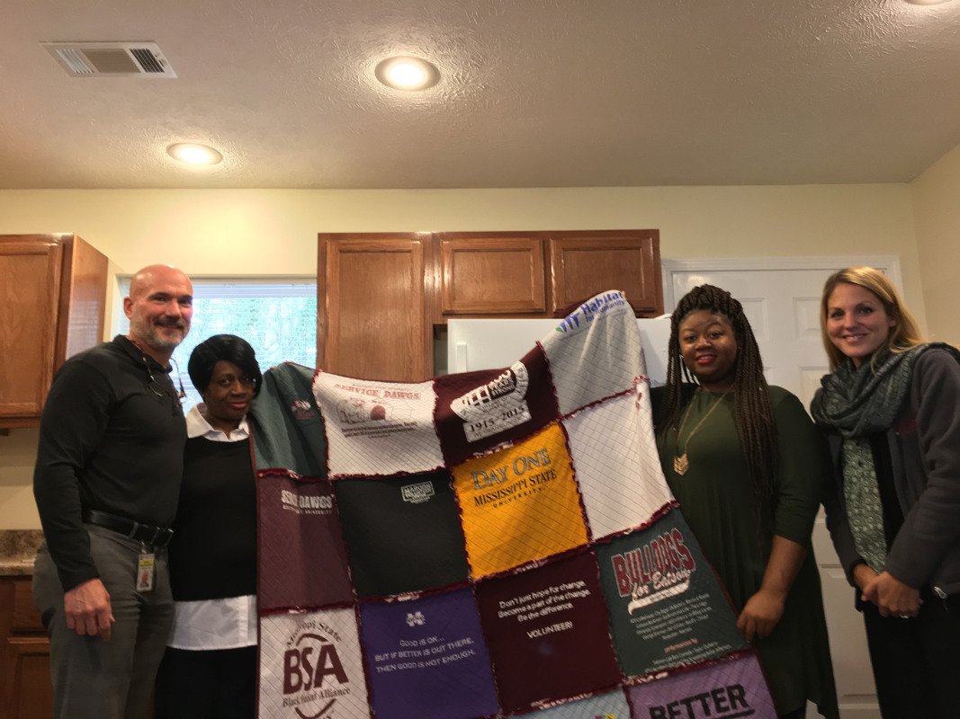 Mississippi State University Director of Student Leadership and Community Engagement Cade Smith (far left) and assistant director Meggan Franks (far right), present a handmade t-shirt quilt to Habitat for Humanity partner family Lena and LaShay Evans. The quilt is designed to help the Evans family remember the MSU students that worked on their home.  (Photo by James Carskadon) 